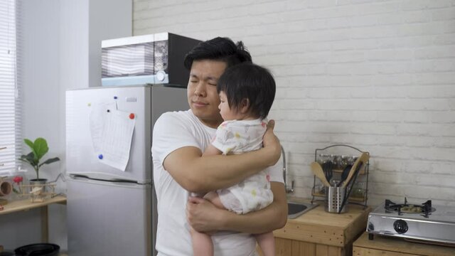 chinese father is shaking his body while singing to his innocent young kid in arms during daytime in the kitchen at home.
