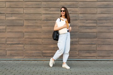 Full length photo shot of young charming beautifyl brunet woman going in the street near brown wall looking to side in white t-shirt and light blue jeans white sneakers with black female bag holding