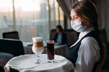 A female Waiter of European appearance in a medical mask serves Latte coffee.