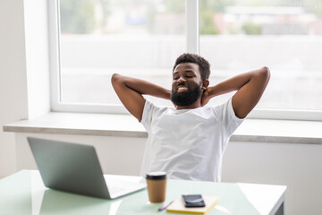 Cheerful african man is watching at his laptop screen, at his work place, with arms behind the...
