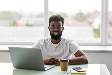 Image of young African businessman with cup of coffee looking at laptop at workplace