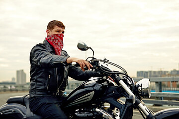 Portrait of stylish male dressed in a black leather jacket and bandana sitting on his custom-made retro motorcycle, looking away, buildings of the big city on background.