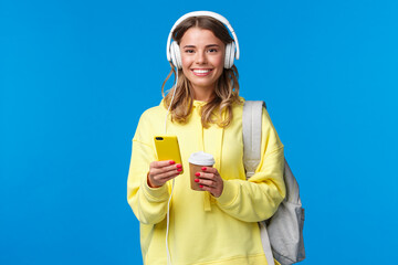 Education, lifestyle and teenagers concept. Cheerful young blond girl in headphones listening music and texting friends while waiting for them near cafe, drinking take-away coffee hold backpack