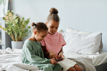 Portrait of two cute African-American girls using smartphone together while sitting on bed in cozy room, copy space