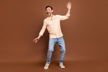 Fototapeta na wymiar Photo of positive funny guy dance have fun wear beige shirt jeans sneakers isolated brown color background