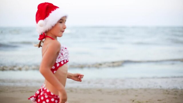 5-year-old girl in red swimsuit and Santa hat on beach is happy, dancing, jumping and running with happiness. Christmas and New Year winter tours to warm countries. Tourism for holidays in tropics