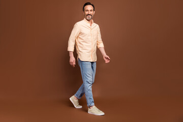 Fototapeta na wymiar Photo of carefree bearded mature guy enjoy walk wear beige shirt jeans shoes isolated brown color background