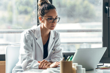 Female doctor talking while explaining medical treatment to patient through a video call with laptop and earphones in the consultation.