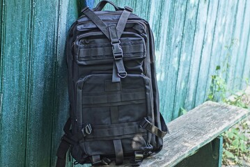 one black tactical backpack stands on a gray wooden bench against a green wall on the street