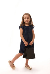 Black Friday concept. Isolated full-length portrait on white background with copy space of an adorable 4 years old child girl dressed in evening attire and golden shoes, holding a shopping packet.