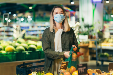 Fototapeta premium Young woman in face medical protective mask chooses and picks in eco bag vegetables or fruits in the supermarket Female standing in a grocery store market near the counter buys in a reusable package