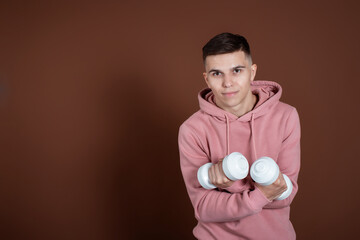 Portrait of a young guy with dumbbells in his hands. Sports lifestyle. The man on a brown background.