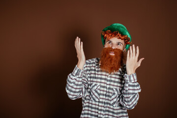 A funny guy in a plaid shirt and a leprechaun hat shows hand gestures. The man on a brown background. Celebration and tradition.