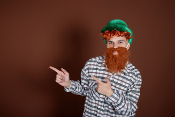 Fototapeta na wymiar A funny guy in a plaid shirt and a leprechaun hat shows hand gestures. The man on a brown background. Celebration and tradition.