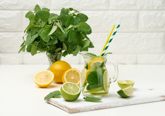 transparent glass decanter with slices of lemon, lime and mint leaves on a white table, detox....