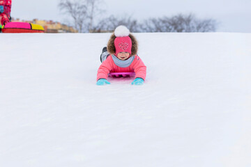 Fototapeta na wymiar Funny little toddler girl in pink jumpsuit sledding down the hill. wintertime for children. winter games outdoors. Kid having fun at Christmas time