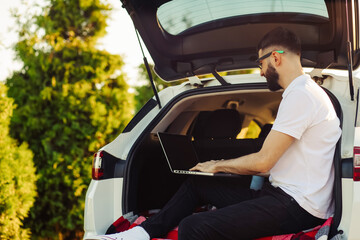 Young man enjoying a trip in his car, sitting in the trunk of a car with a laptop