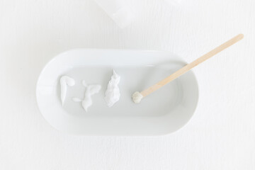 an oval saucer with smears of cosmetic cream. a wooden stick with cream is lying next to it. there are several creamy textures on a white background. Cosmetology and care.