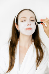 caucasian girl in a white coat with a white mask on her face. the cosmetologist applies the mask to the face with a brush. Facial peeling mask, spa beauty treatments, skin care.