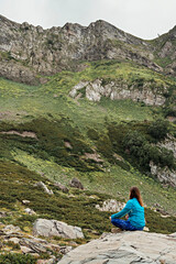Fototapeta premium rear view of meditating young woman in blue sportswear sitting on stone in green mountain valley, relaxation in nature, consciousness, mindfulness