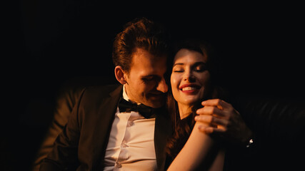 happy man in suit hugging and flirting with cheerful girlfriend isolated on black