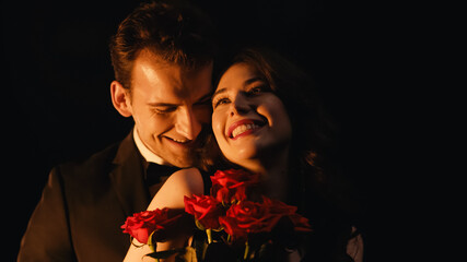cheerful woman receiving red roses from man standing behind isolated on black