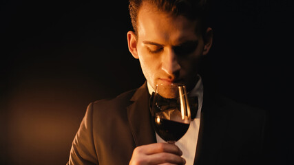 Confident young man in suit holding glass and smelling red wine on black