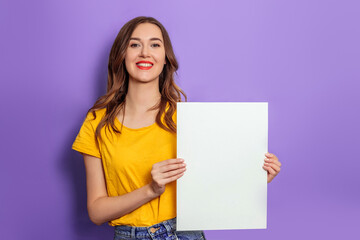 Fototapeta na wymiar Caucasian young woman smiling and holding blank poster wearing yellow t-shirt isolated over lilac background in studio. Mockup for design