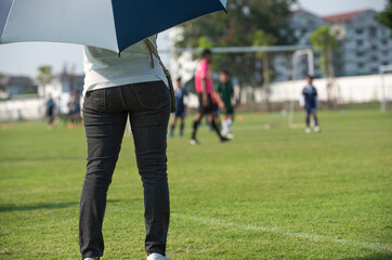 Fototapeta premium Mother standing and watching her son playing football in a school tournament on a clear sky and sunny day. Sport, active lifestyle, happy family and soccer mom concept.