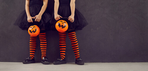Halloween party banner. Cropped image of legs of two little girls with pumpkin candy bucket standing on black background. Children in black dresses and black and orange leggings stand near copy space