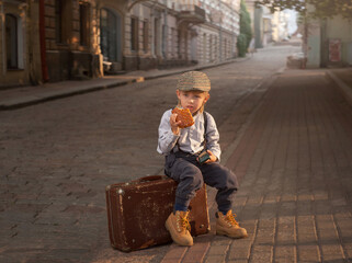 a boy in vintage clothes is sitting on a suitcase by the road and eating a bun. He is wearing a...