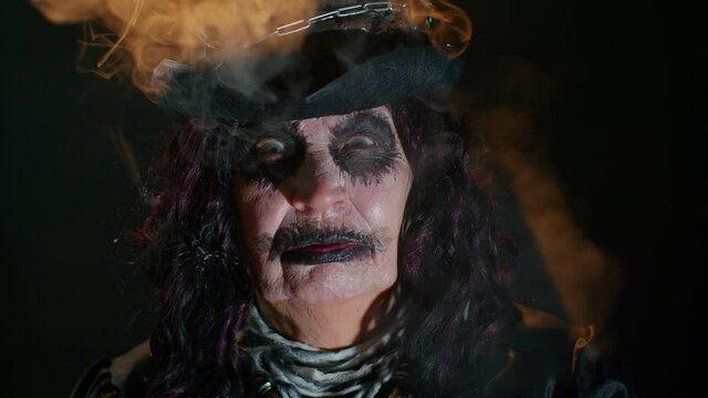 Creepy mature woman grandmother with Halloween stylish witch makeup. Old scary granny making faces, blows smoke from mouth, trying to scare. Voodoo ritual. Thematic party. Sinister senior lady vampire