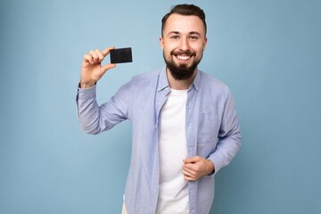 Closeup photo of Good looking cool smiling brunette bearded young man wearing stylish blue shirt and white t-shirt isolated over blue background wall holding credit card looking at camera