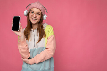 Beautiful happy smiling young woman wearing stylish casual outfit isolated on background wall holding and showing mobile phone with empty display for mockup wearing white bluetooth headphones