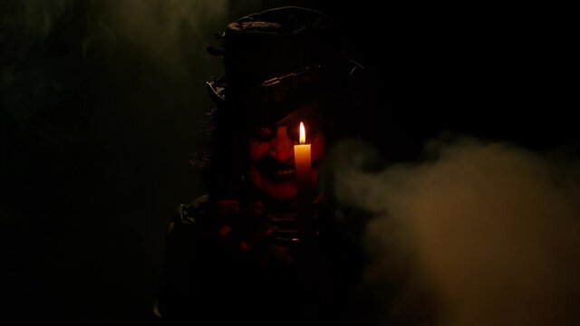 Sinister old elderly woman with horrible scary Halloween witch makeup in costume looking ominous at camera, making voodoo magic rituals with candle. Vampire granny isolated against black background