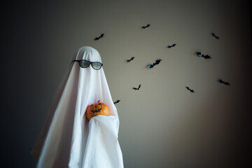 Ghost from sheet with glasses stands against background of bats and hold pumpkin. Happy halloween