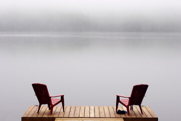 two Adirondack chairs on wooden deck at edge of lake in fog with copy space