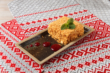Piece of cake Napoleon on brown plate on textile with national belorussian ornament.