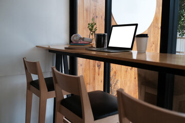 Photo of an empty screen computer laptop putting on wooden counter bar surrounded by a takeaway coffee cup, a cup of coffee beans, headphones, a stack of books, and a potted plant.