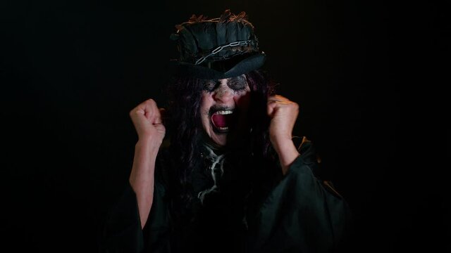 Frustrated sinister woman with horrible scary Halloween witch makeup raising hands in indignant expression, quarreling, asking reason of conflict, why such stubborn. Vampire granny on black background