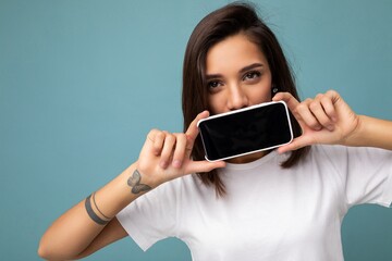 closeup Photo of pretty positive young brunette woman good looking wearing white t-shirt standing isolated on blue background with copy space holding smartphone showing phone in hand with empty screen