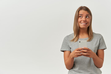Indoor shot of young adult female, wears gray t-shirt, texting with her boyfriend, looks aside and biting her lip, smiles broadly. Isolated over white background