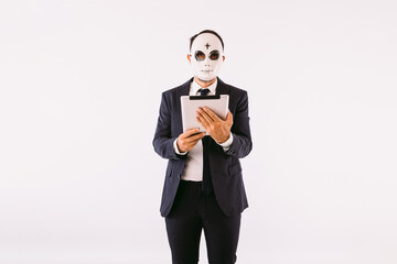Man dressed in suit and tie, wearing a killer's mask with a cross on his forehead for Halloween, looking at his tablet. Carnival and halloween celebration