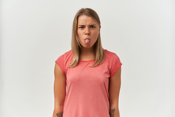 Young adult female, shows her tongue with negative, upset facial expression. Isolated over white...