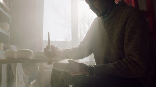 Young man carpenter is sitting near window and cleaning wooden item, working with inspiration in workshop, Soft Counter Light, Close up, Slow motion.