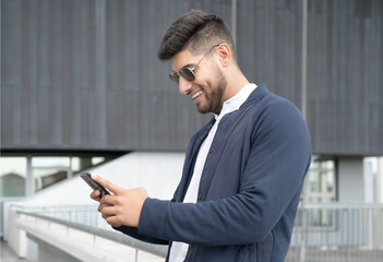 Handsome man using mobile phone app texting outside of office in urban city with skyscrapers buildings in the background. Young man holding smartphone for business work. High quality photo