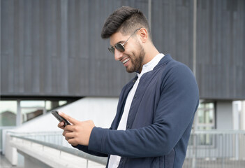 Fototapeta premium Handsome man using mobile phone app texting outside of office in urban city with skyscrapers buildings in the background. Young man holding smartphone for business work. High quality photo