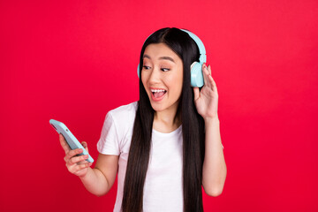 Photo of young asian girl happy smile listen music earphones look browse smartphone isolated over red color background