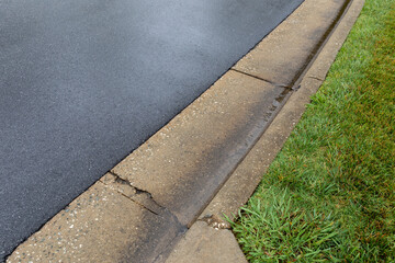 Diagonal view of a wet asphalt roadway draining into a formed concrete curb with grassy...