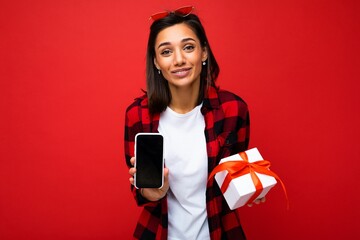 Photo of beautiful positive surprised young brunette woman isolated over red background wall wearing white casual t-shirt and red and black shirt holding white gift box with red ribbon and mobile